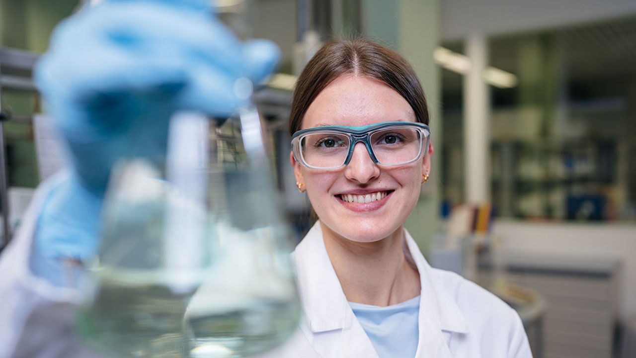 Person im Labor mit Schutzbrille und Kolben in der Hand