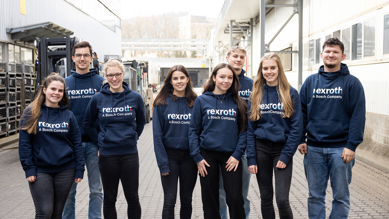 Gruppenbild mit 8 jungen Menschen, die dunkelblaue Hoodies der Bosch Rexroth AG tragen und in einem Außenbereich zwischen zwei Firmengebäuden stehen. Alle schauen fröhlich.