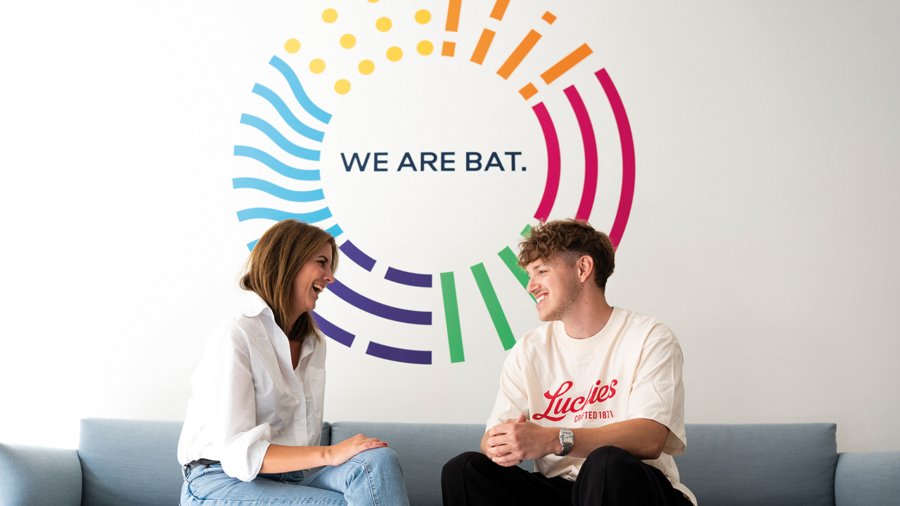 Zwei junge Menschen sitzen auf einem Sofa und unterhalten sich. Im Hintergrund ein buntes Bild mit dem Slogan "We are BAT."