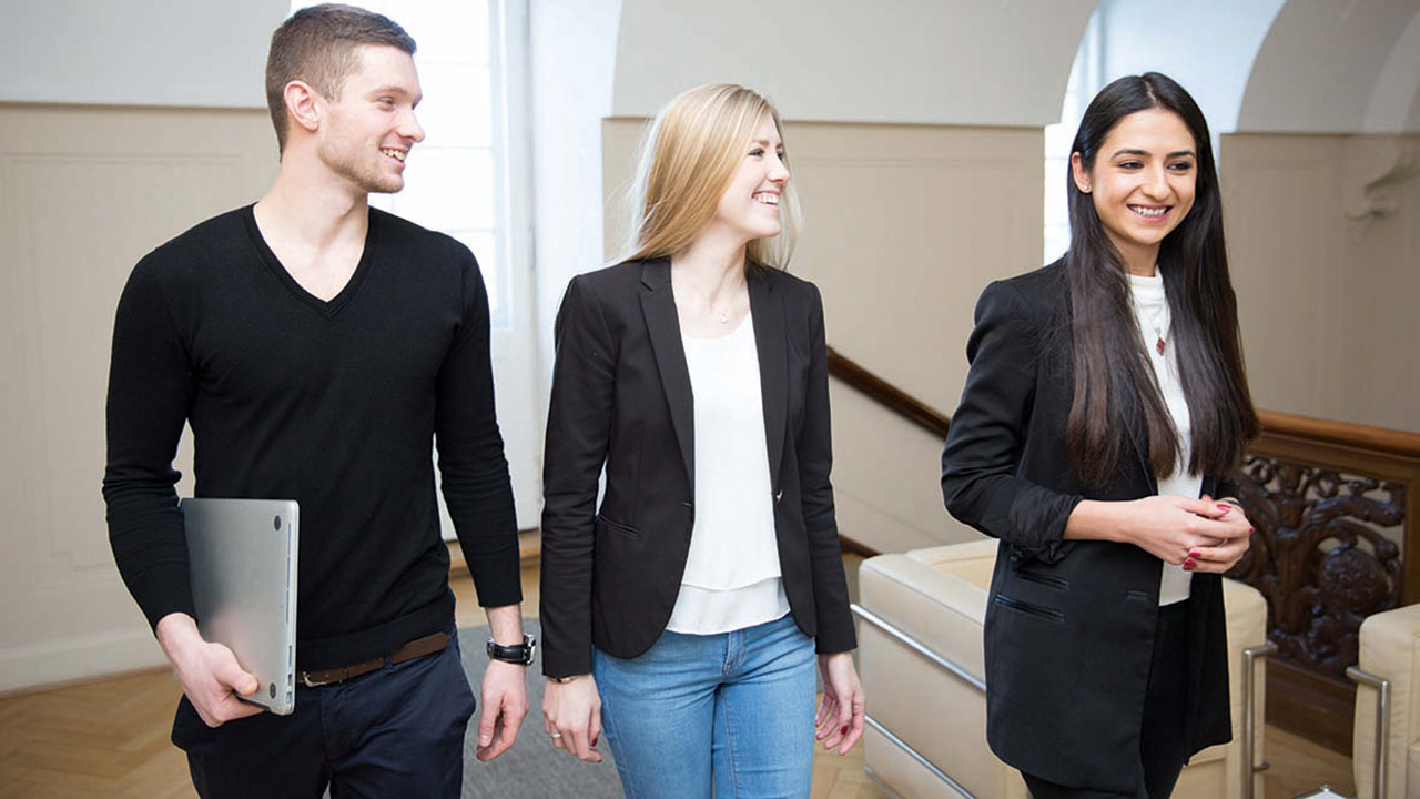 Drei junge lächelnde Personen in Businesskleidung, eine davon mit Laptop unter dem Arm