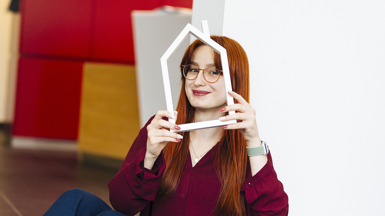 Frau hält einen Bilderrahmen in Form eines Hauses vor sich und lacht in die Kamera