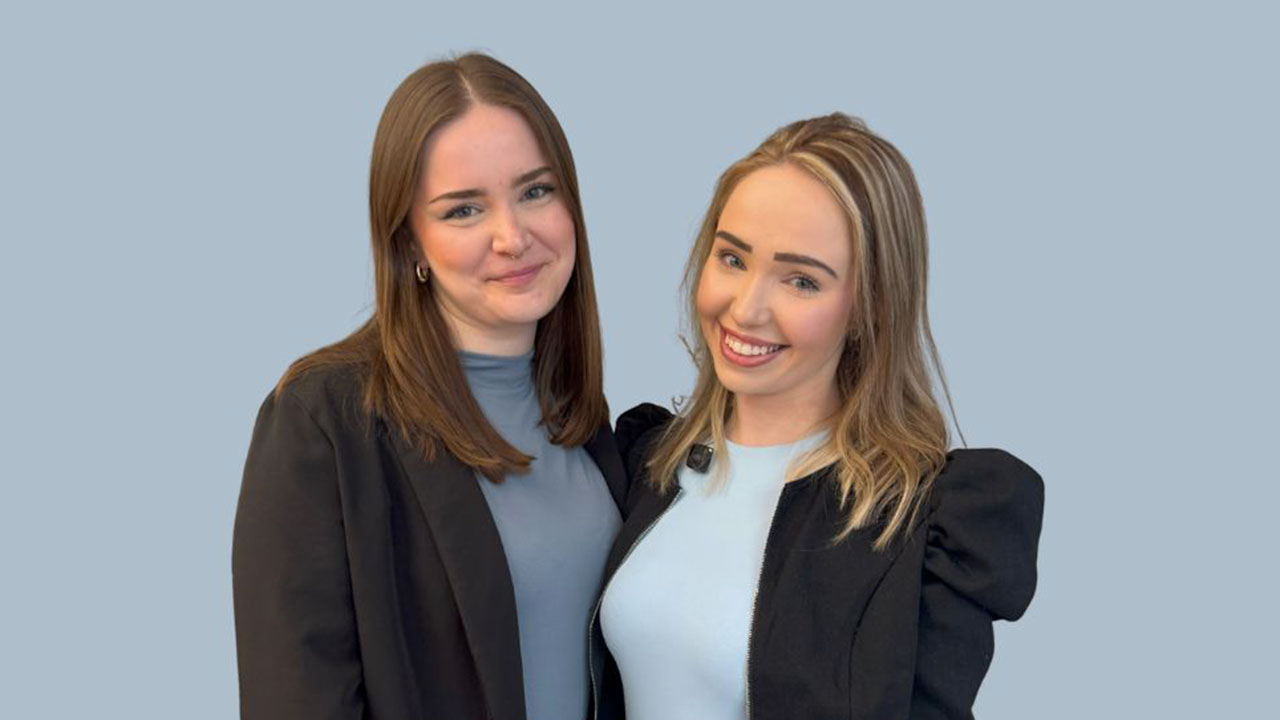 Zwei junge Personen mit mittlellangen Haaren lächeln in die Kamera