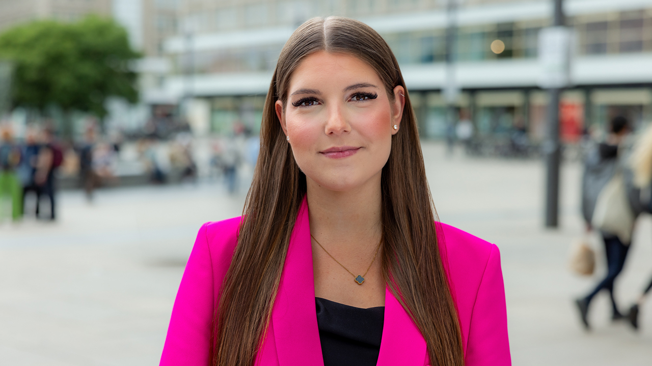 Junge Frau mit pinkem Blazer und langen braunen Haaren, die in die Kamera lächelt