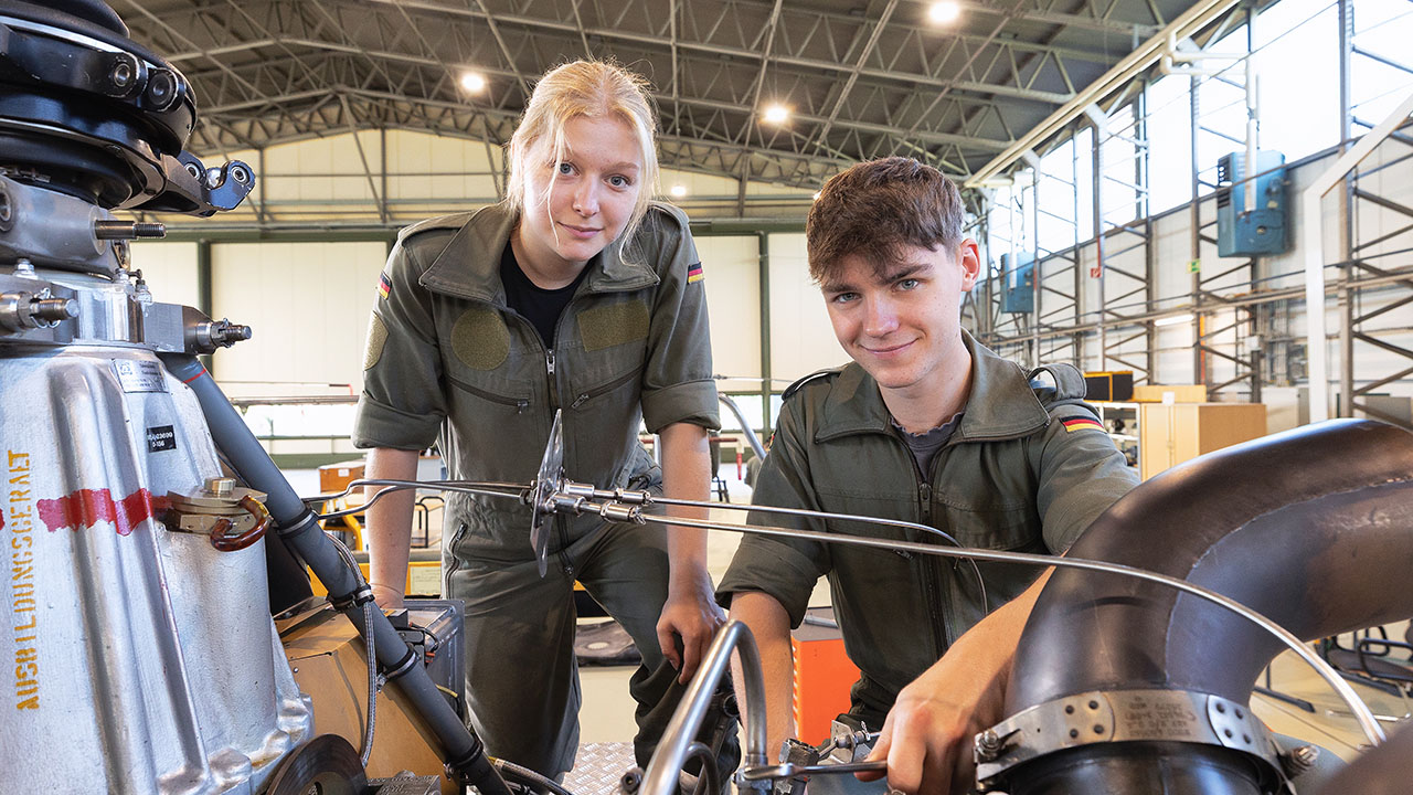 Zwei Jugendliche in dunkelgrüner Bundeswehr-Arbeitskleidung arbeiten an einem Gerät in einer großer Werkstatthalle.
