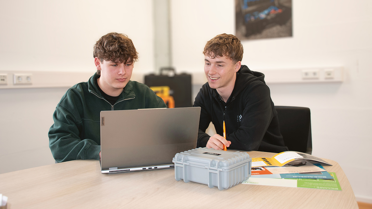Zwei Auszubildende sitzen mit Material und einem Laptop an einem Tisch und besprechen sich.
