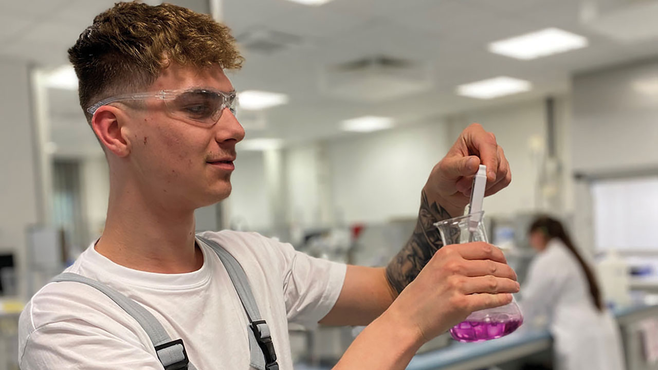 Person hält ein Reagenzglas in der Hand in dem sich lilafarbene Flüssigkeit befindet.