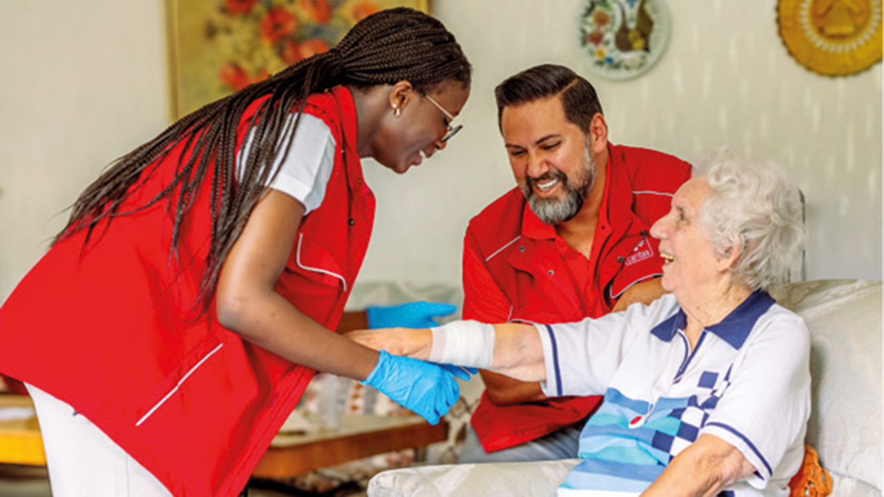 Zwei Personen in roter Pflegebekleidung kontrollieren den Verband am Arm einer Seniorin