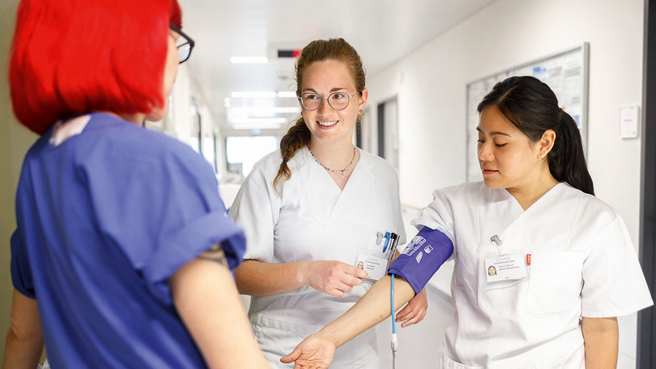 Junge lächelnde Person in weißer Pflegebekleidung zeigt einer Person in blauer Pflegebekleidung, wie die Manschette zum Blutdruckmessen angelegt wird