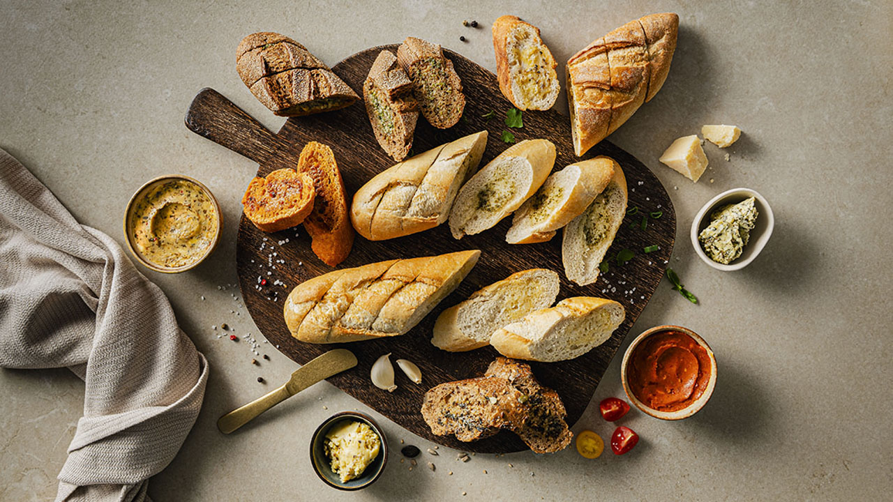 Holzbrett mit Kräuterbaguette und verschiedenen Aufstrichen in kleinen Töpfchen