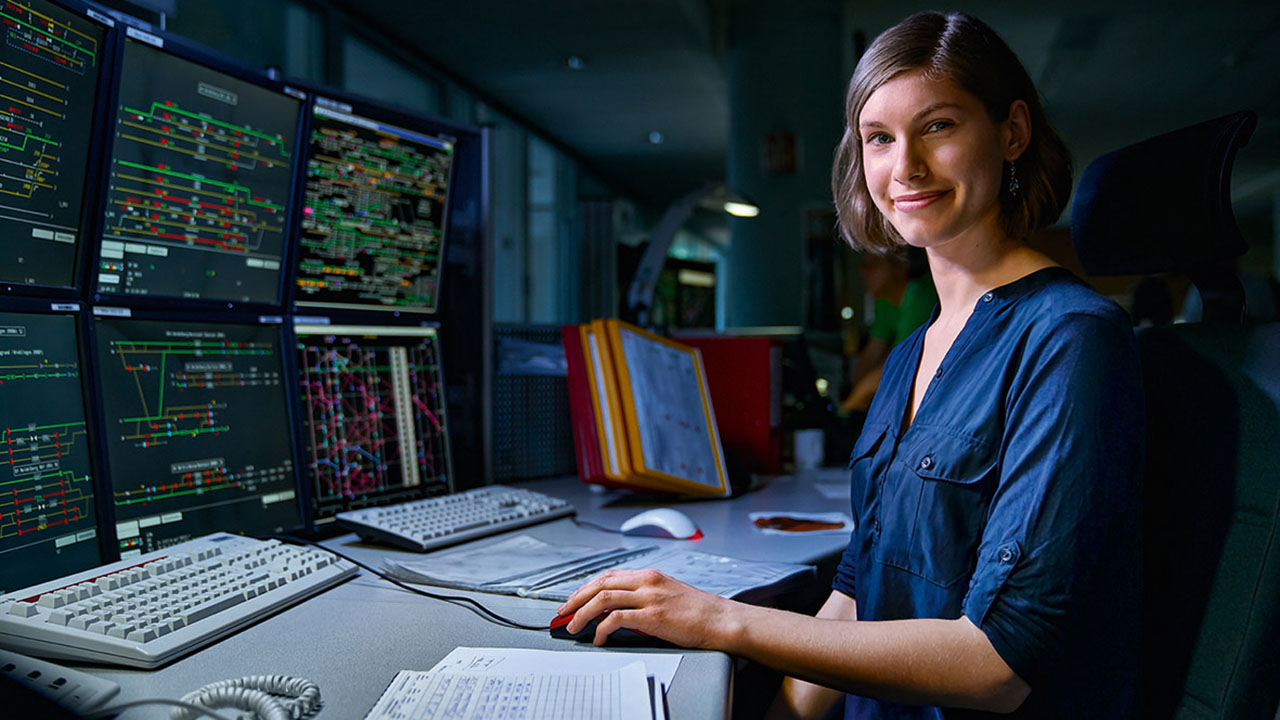 Junge lächelnde Person sitzt an einem Schreibtisch mit sechs Monitoren in der Zugverkehrssteuerung