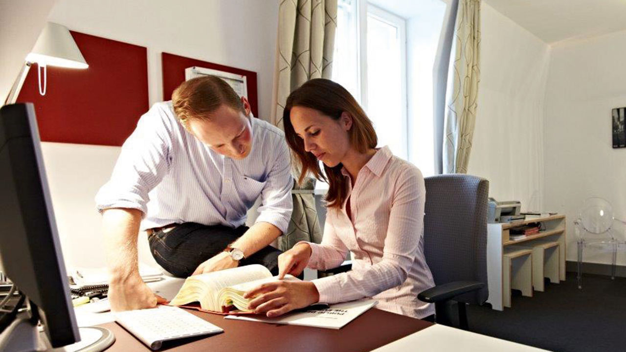 Junge Person sitzt am Schreibtisch und zeigt einer anderen Person einen Passus in einem Buch