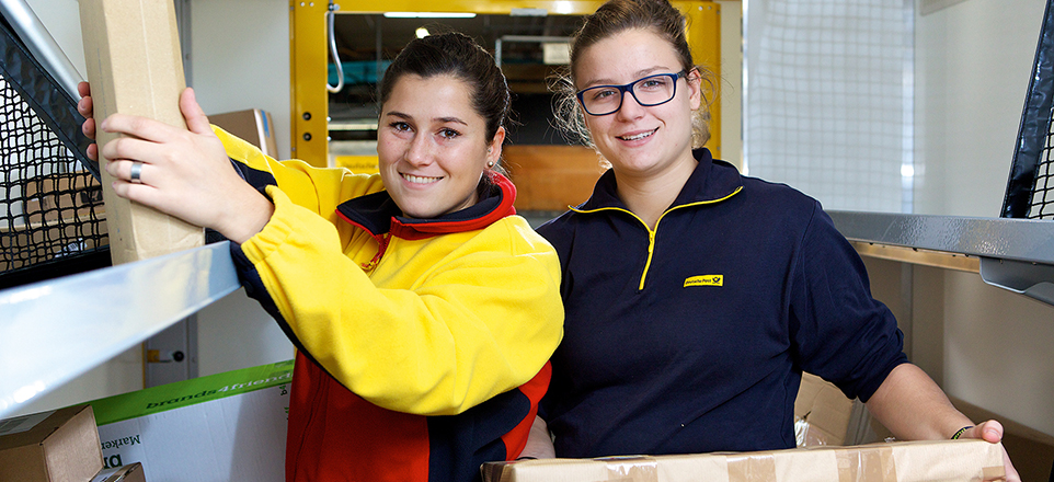 2 junge Frauen mit Post-Arbeitskleidung sortieren Pakete in ein Regal ein und lächeln
