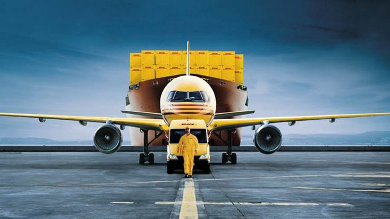 Person in gelber Arbeitskleidung vor einem Transporter, einem Flugzeug und einem Containerschiff