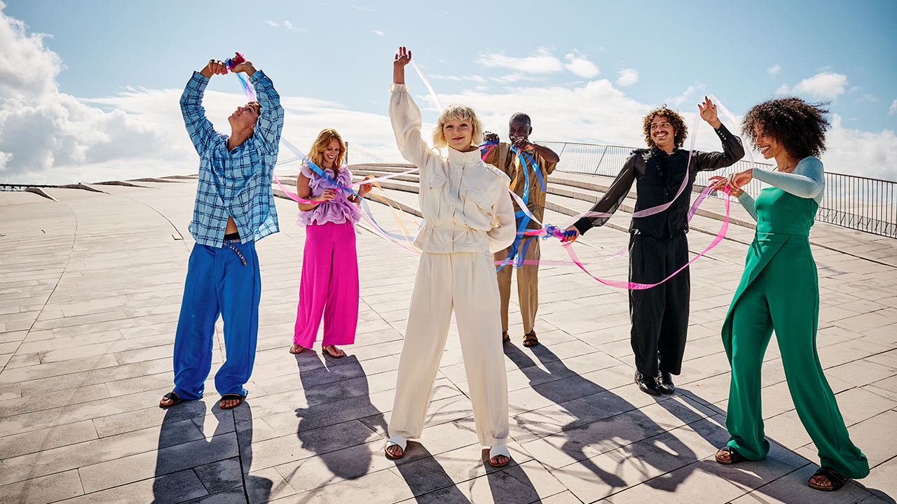 Gruppe junger bunt gekleideter Menschen auf einer Dachterrasse. Alle halten einen Teil eines miteinander verknoteten bunten Bandes.