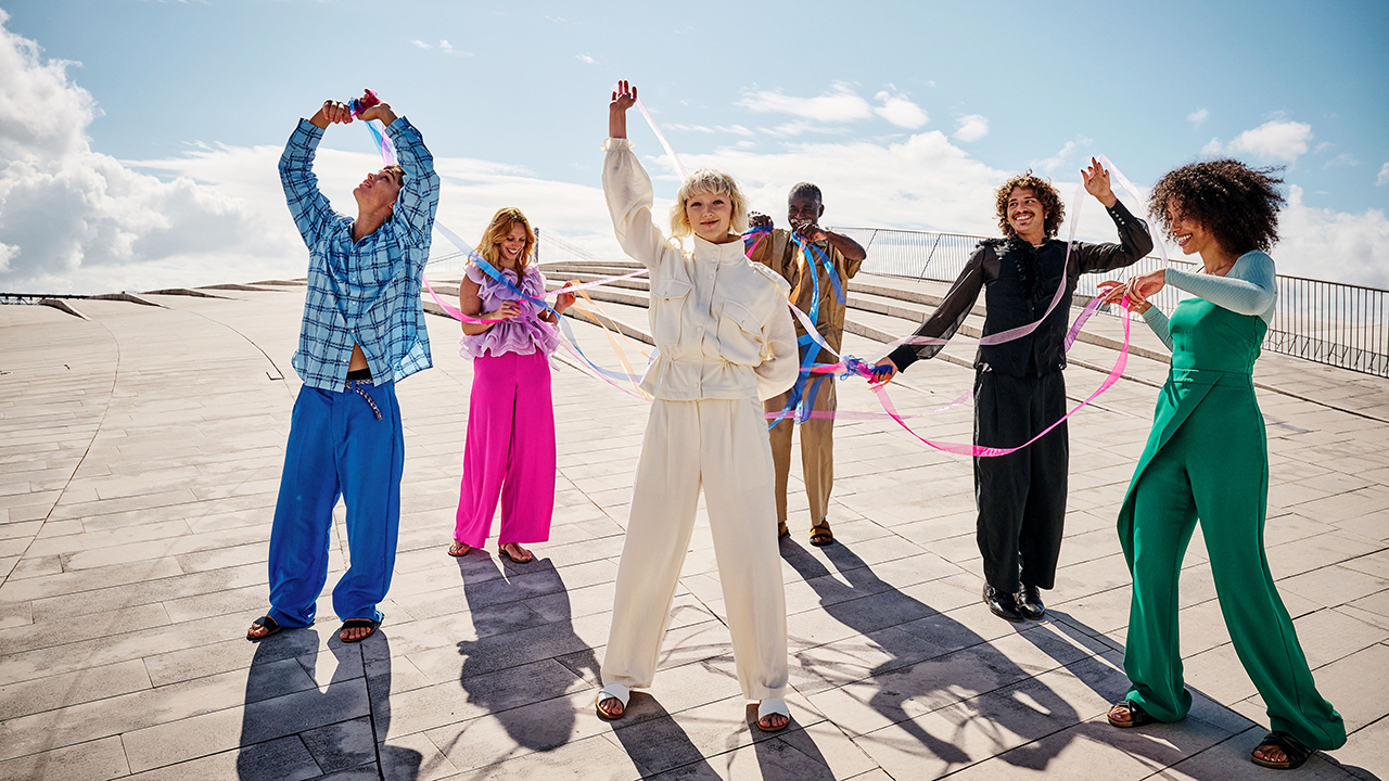 Aufnahme von 6 jungen Menschen auf einer Dachterrasse. Im Hintergrund der blaue Himmel und ein paar vereinzelte Wolken. Die jungen Menschen haben jeweils ein farbiges Outfit an: blau, pink, weiß, gelb, schwarz und grün und halten farbige Bänder, die miteinander verwoben sind.