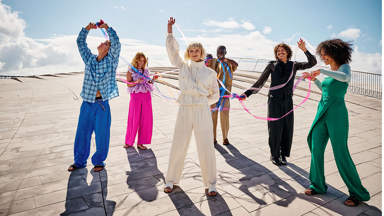 Sechs junge Menschen tanzen in bunten Klamotten und mit bunten Bändern. Die Sonne strahlt sie an und sie werfen Schatten. Der Himmel ist hellblau