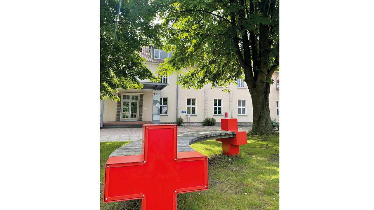 Gebäude mit Bäumen von außen zu sehen- Davor ist eine holzbank mit den typischen roten Kreuz Kreuzen als Bankhalter