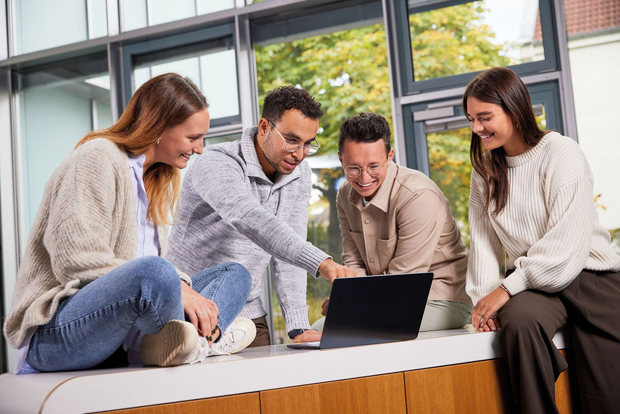 Mehrere Personen sitzen bzw. stehen gemeinsam an einem Tisch und schauen lächelnd auf einen Laptop.