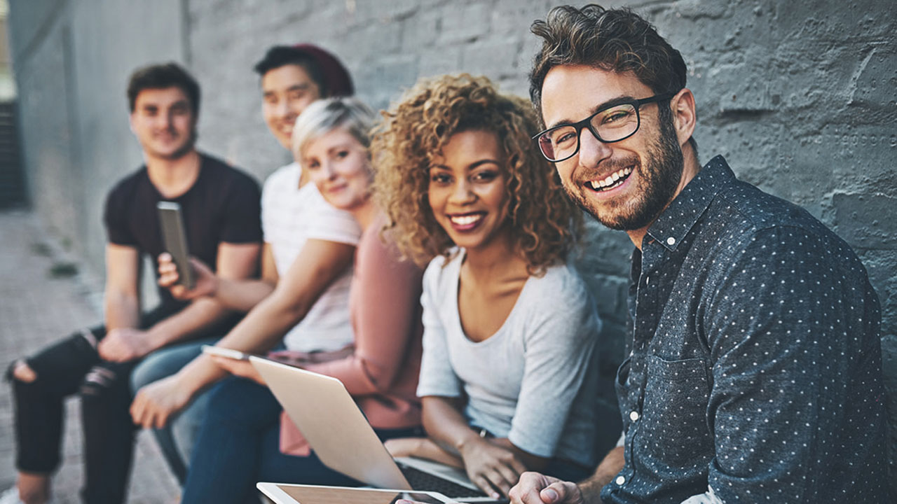 Fünf junge Personen mit Handy, Tablet und Laptop vor einer grauen Mauer lächeln in die Kamera