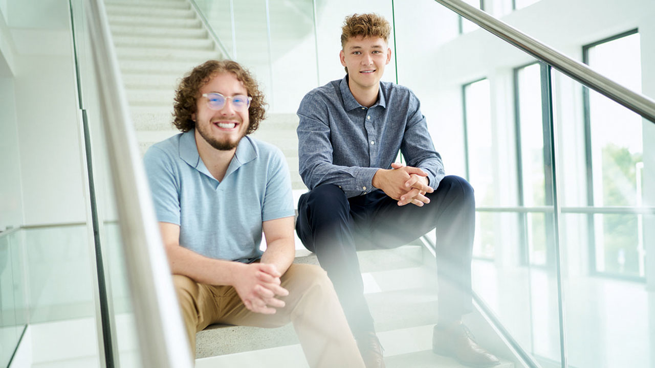 Zwei junge Menschen sitzen auf einer Treppe im Gebäude und lächeln in die Kamera
