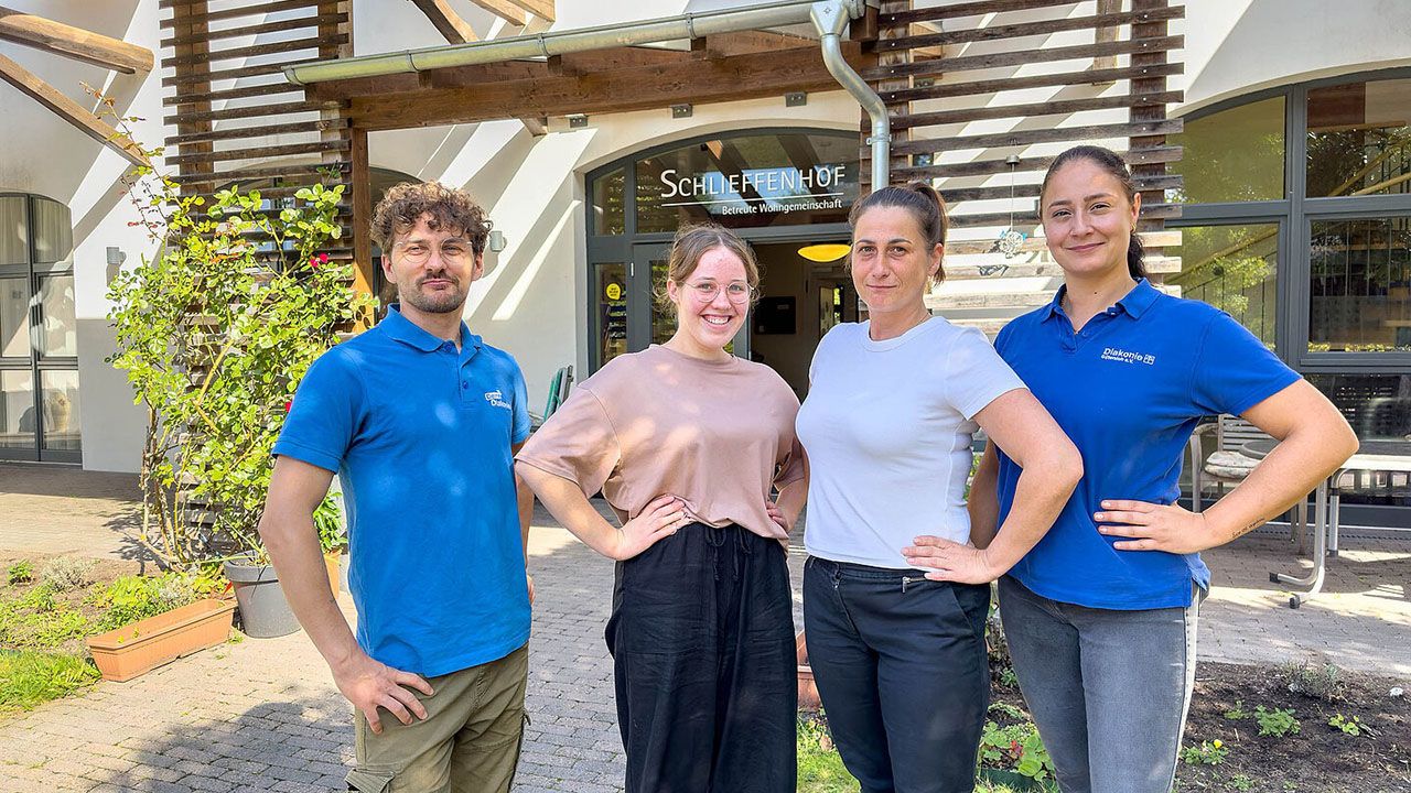 Auf dem Bild stehen vier Personen nebeneinander vor dem Eingang eines Gebäudes. Über der Tür ist ein Schild mit der Aufschrift „Schlieffenhof – Betreute Wohngemeinschaft“.