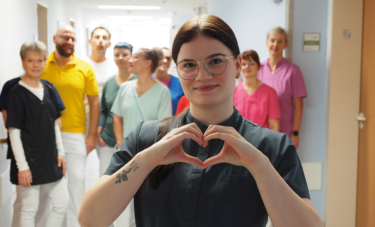 Gruppenbild in einem Krankenhausflur, ganz vorn steht eine junge Person und formt aus ihren Fingern ein Herz.
