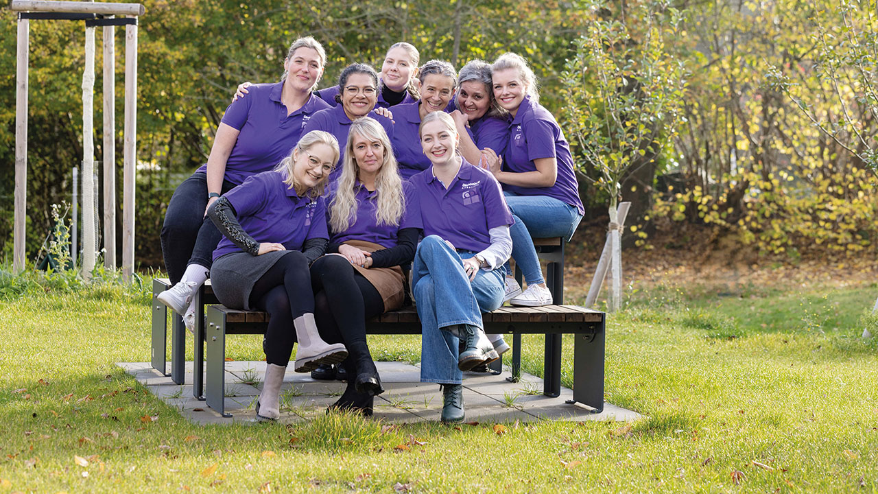 Eine Gruppe von lächelnden Personen sitzt eng beieinander auf einer Bank im Grünen, alle tragen lila Oberteile.