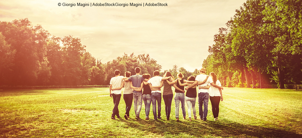 Gruppe von zehn jungen Menschen läuft Arm in Arm über eine Wiese, die von Bäumen gesäumt ist