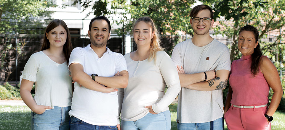 5 junge Menschen lächeln freundlich in die Kamera. Sie sind im Halbkörperporträt dargestellt und stehen bei schönem Wetter draußen eng zusammen.