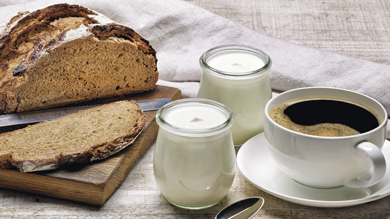 Stillleben - Brett mit Brotmesser und aufgeschnittenem Brot dazu eine Tasse Kaffe und zwei Gläser Joghurt.