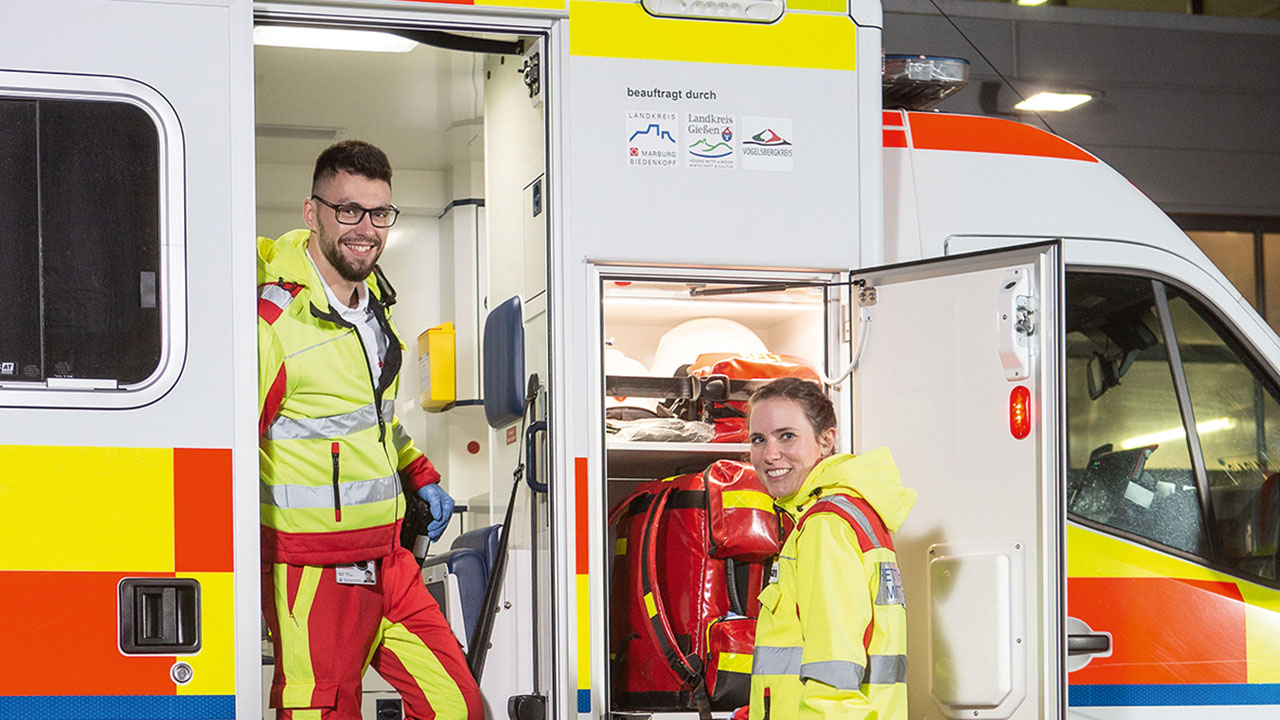 Zwei Personen in leuchtender Schutzkleidung stehen lachend an einem geöffneten Rettungswagen. Eine Person steht im Türrahmen, die andere Person steht davor mit Blick zur Kamera.
