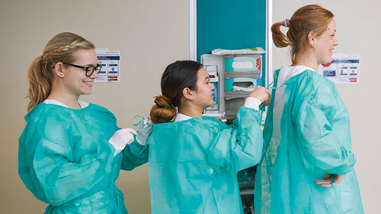 Drei junge Personen stehen hintereinander und binden sich gegenseitig den OP-Kittel zu