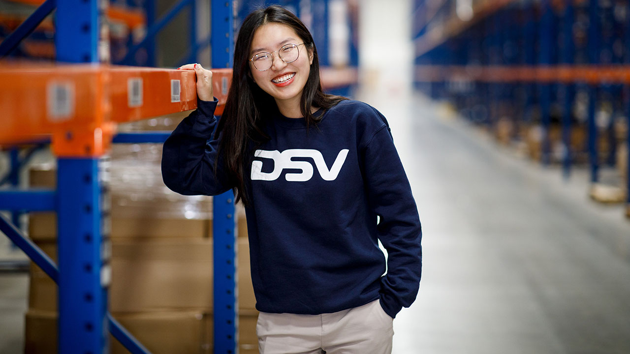 Junge lächelnde Person mit Brille und Pullover mit Firmenlogo in einer Lagerhalle