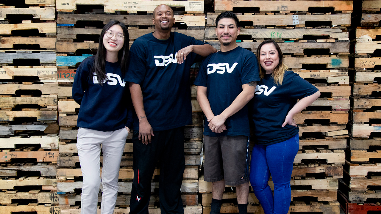 4 junge Menschen in DSV T-shirts vor einer Wand aus Holzpaletten, alle sind gut gelaunt.