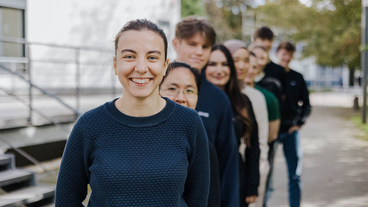 Gruppenfoto von jungen Menschen im Freien, die hintereinander in einer Reihe stehen und in die Kamera lächeln