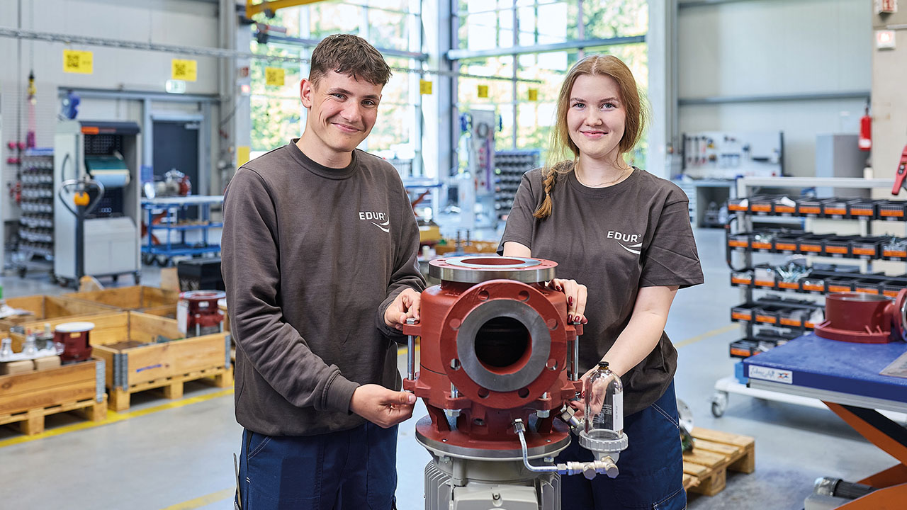 Zwei junge lächelnde Mitarbeitende arbeiten an einer Vorrichtung in der Produktionsstätte