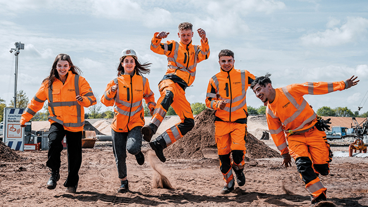 5 junge Menschen in orangener Arbeitskleidung springen auf einer Baustelle in die Luft