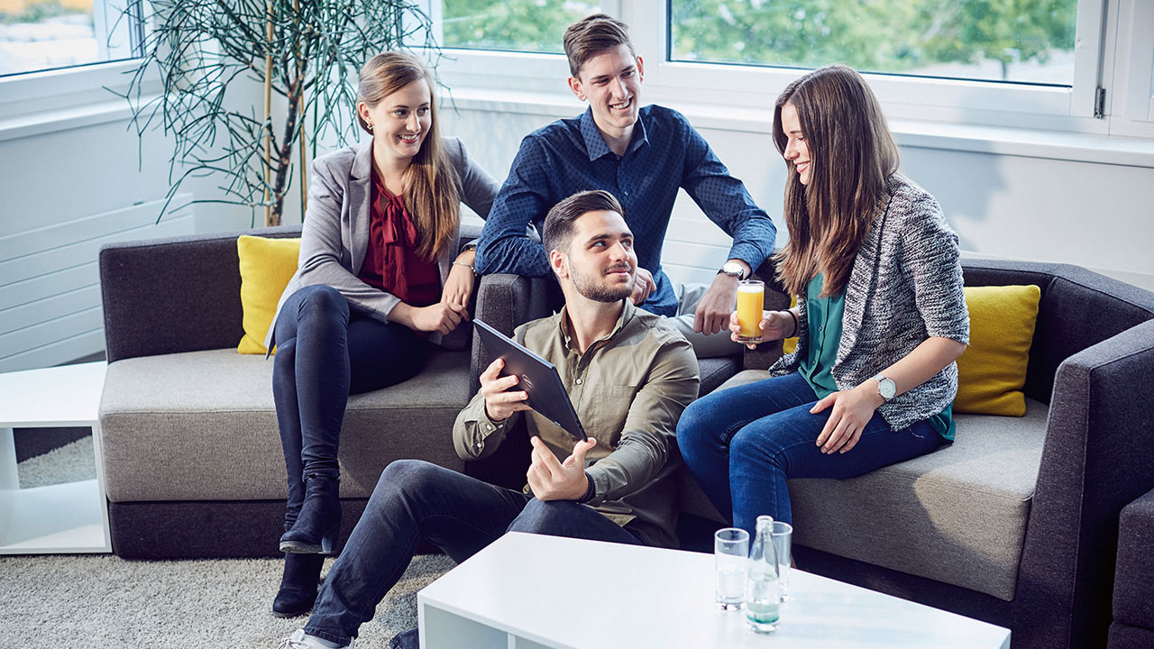 Drei junge Personen sitzen auf einem Sofa, eine Person sitzt vor dem Sofa und zeigt etwas auf einem Tablet