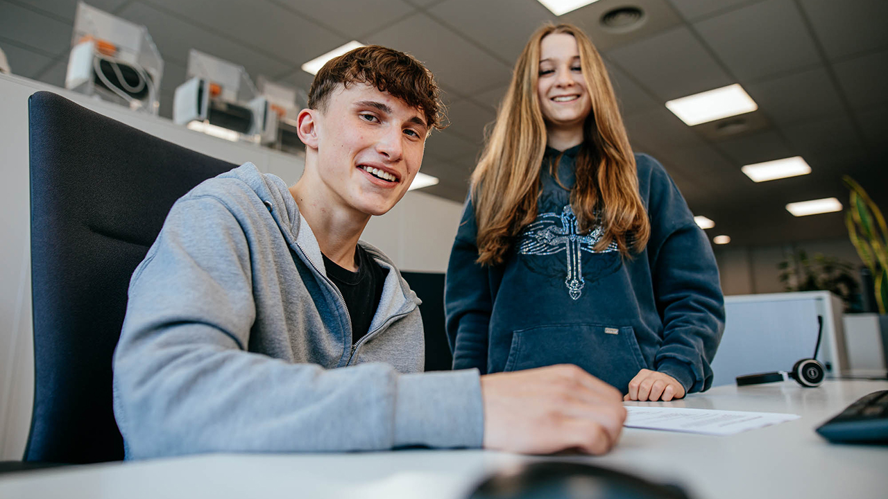 Zwei junge Menschen sitzen an einem Schreibtisch und lächeln in die Kamera