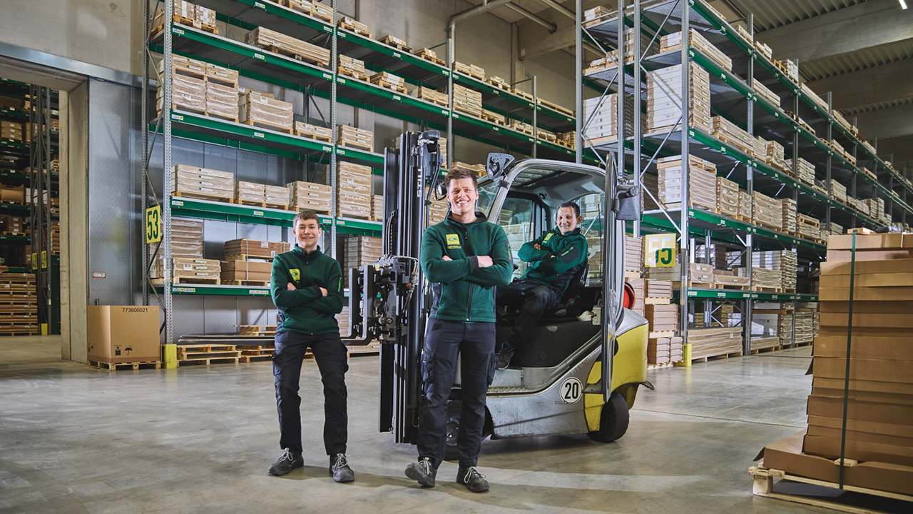 3 junge Mitarbeitende in einer Lagerhalle. Einer von Ihnen sitzt in einem Gabelstapler.