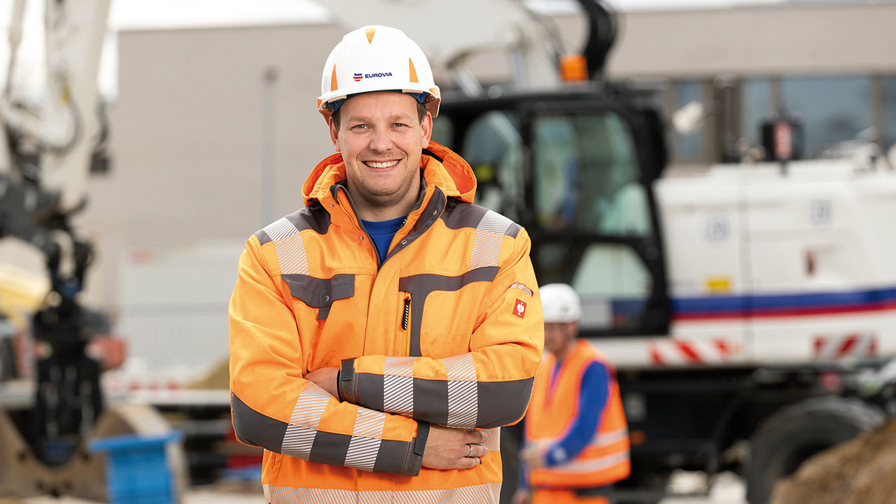 Person in Arbeitskleidung und Arbeitshelm blickt in die Kamera