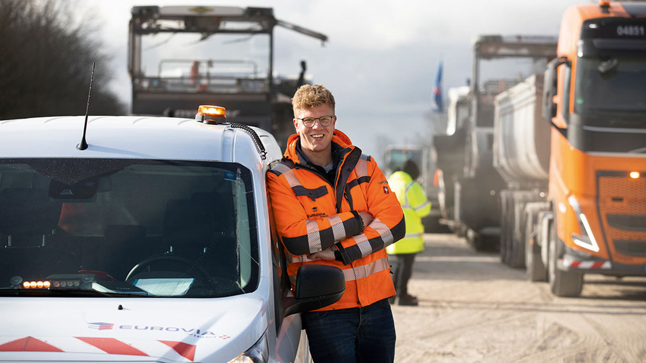 Junge lächelnde Person in orangefarbener Jacke lehnt an einem EUROVIA-Wagen, im Hintergrund Baustellenfahrzeuge