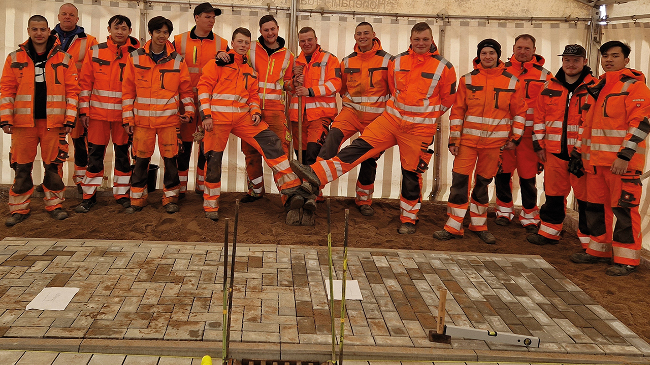 Das Bild zeigt eine Gruppe von Auszubildenden in Arbeitskleidung hinter einer gepflasterten Fläche. 