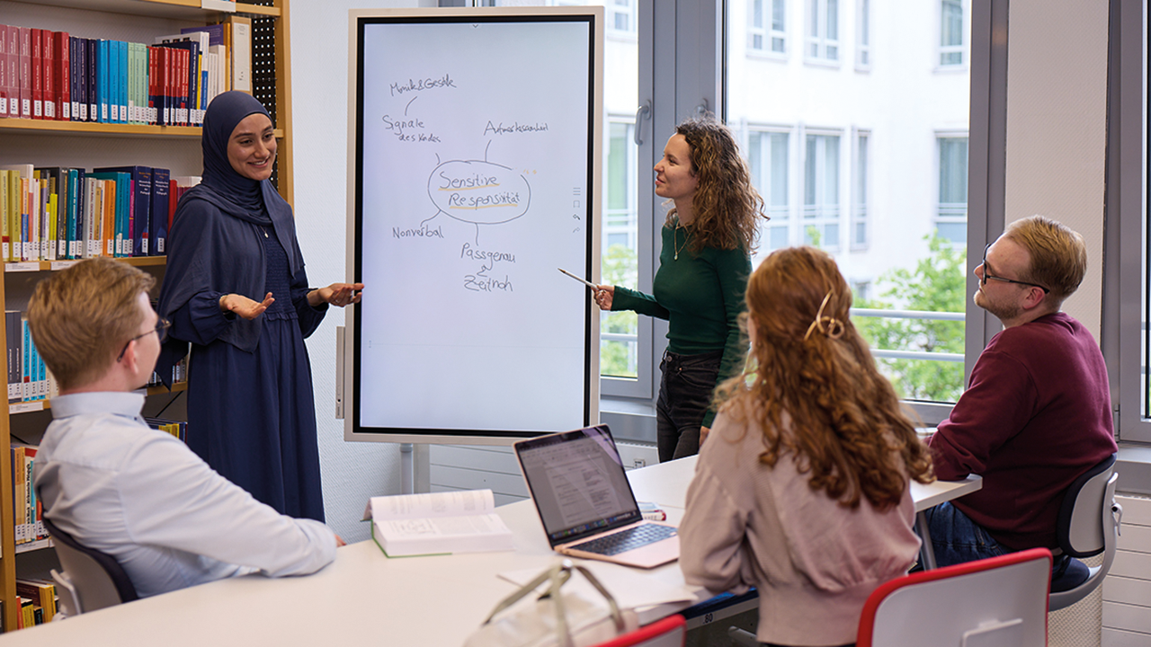 Situation in einem Seminarraum. 3 sitzende Personen die zwei weiteren Personen zuhören, die an einem Flipchart etwas zeigen.