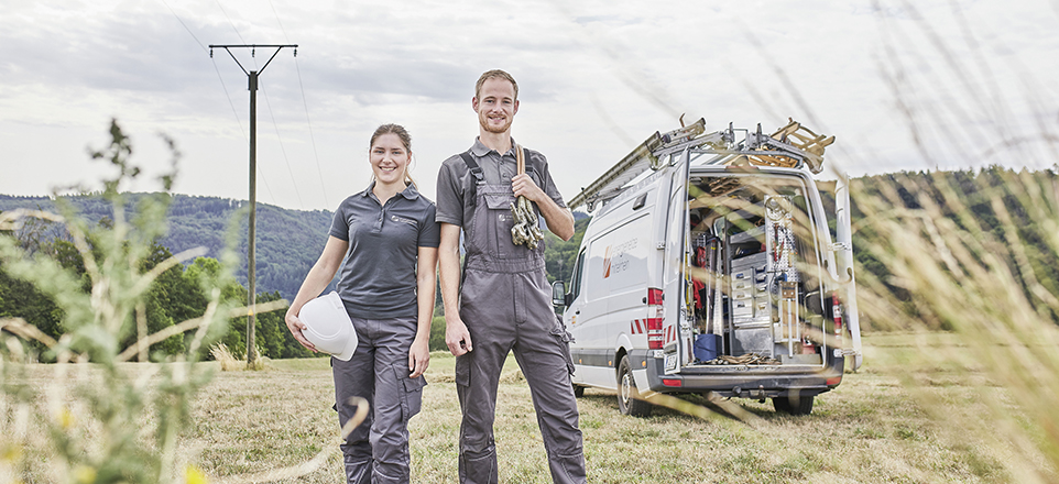 2 Personen in grauen Arbeitsanzügen stehen in der Natur, im Hintergrund stehen ein weißes Auto und ein Strommast