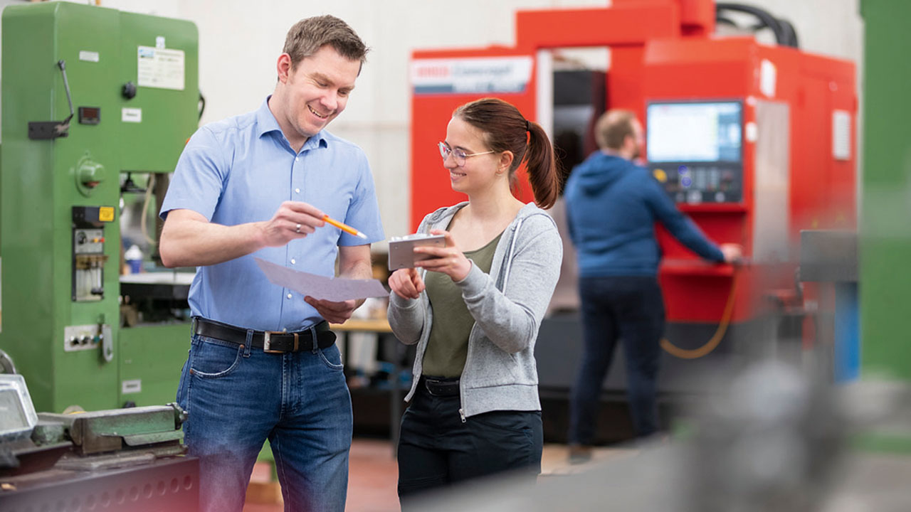 Eine erwachsene Person erklärt einer jüngeren Person etwas, im Hintergrund steht eine Person an einer Maschine