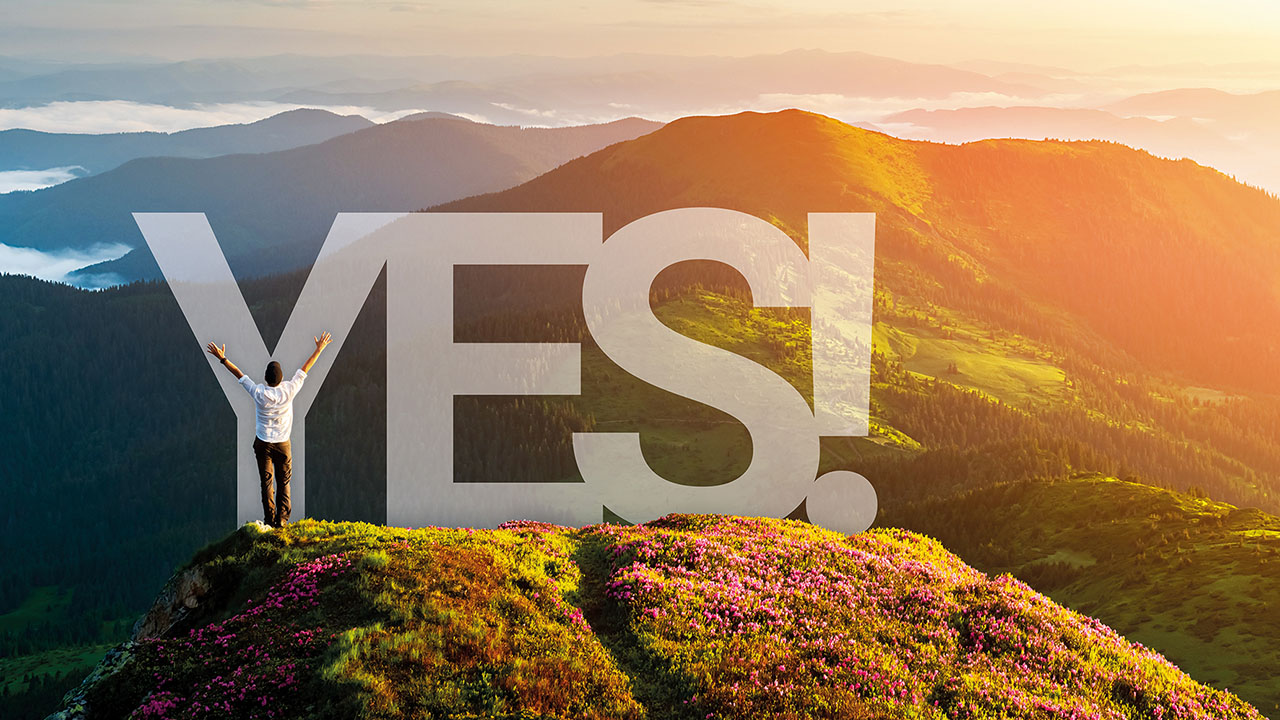 Eine Person steht mit erhobenen Armen auf einem blühenden Berggipfel. Im Hintergrund sind Berge im Morgenlicht zu sehen. Groß steht das Wort „YES!“ im Bild.
