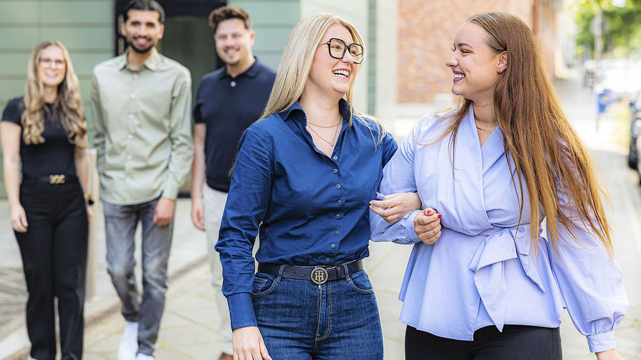 Im Vordergrund zwei junge lächelnde Menschen, die sich unterhaken, im Hintergrund drei weitere junge Menschen