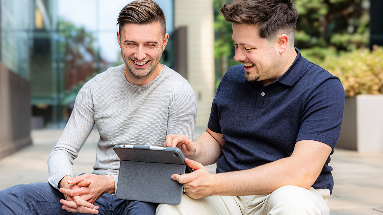 Zwei junge Männer sitzen draußen und schauen in einem Tablet etwas nach