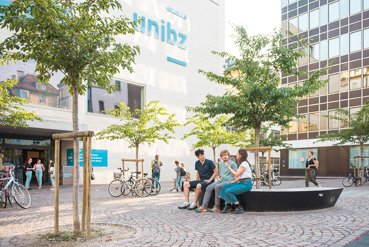 Das Bild zeigt einen Platz vor einem modernen Uni-Gebäude mit dem Schriftzug "unibz". Mehrere Personen sitzen oder stehen, einige unterhalten sich oder stehen bei Fahrrädern.